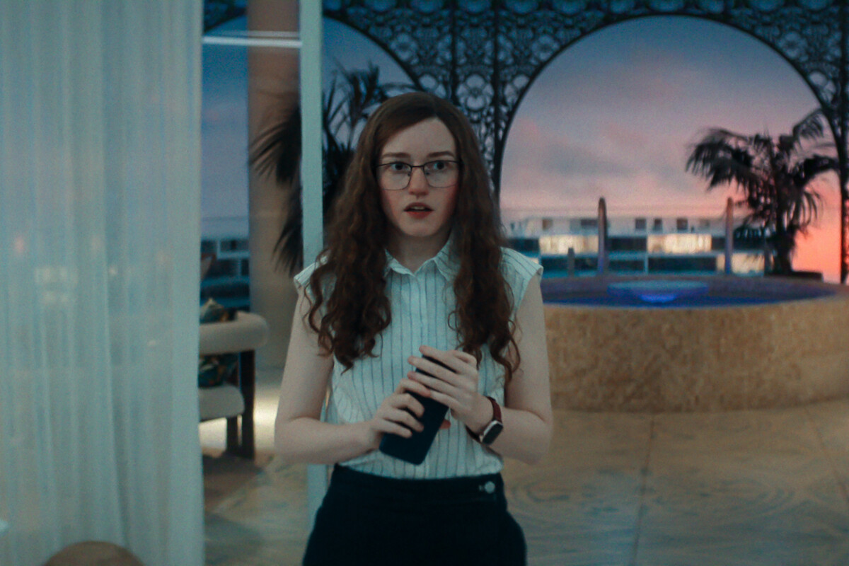 Woman with long curly hair and glasses stands indoors near a decorative pool at sunset, holding a phone, with palm trees and ornate arches in the background, creating a modern, upscale ambiance.