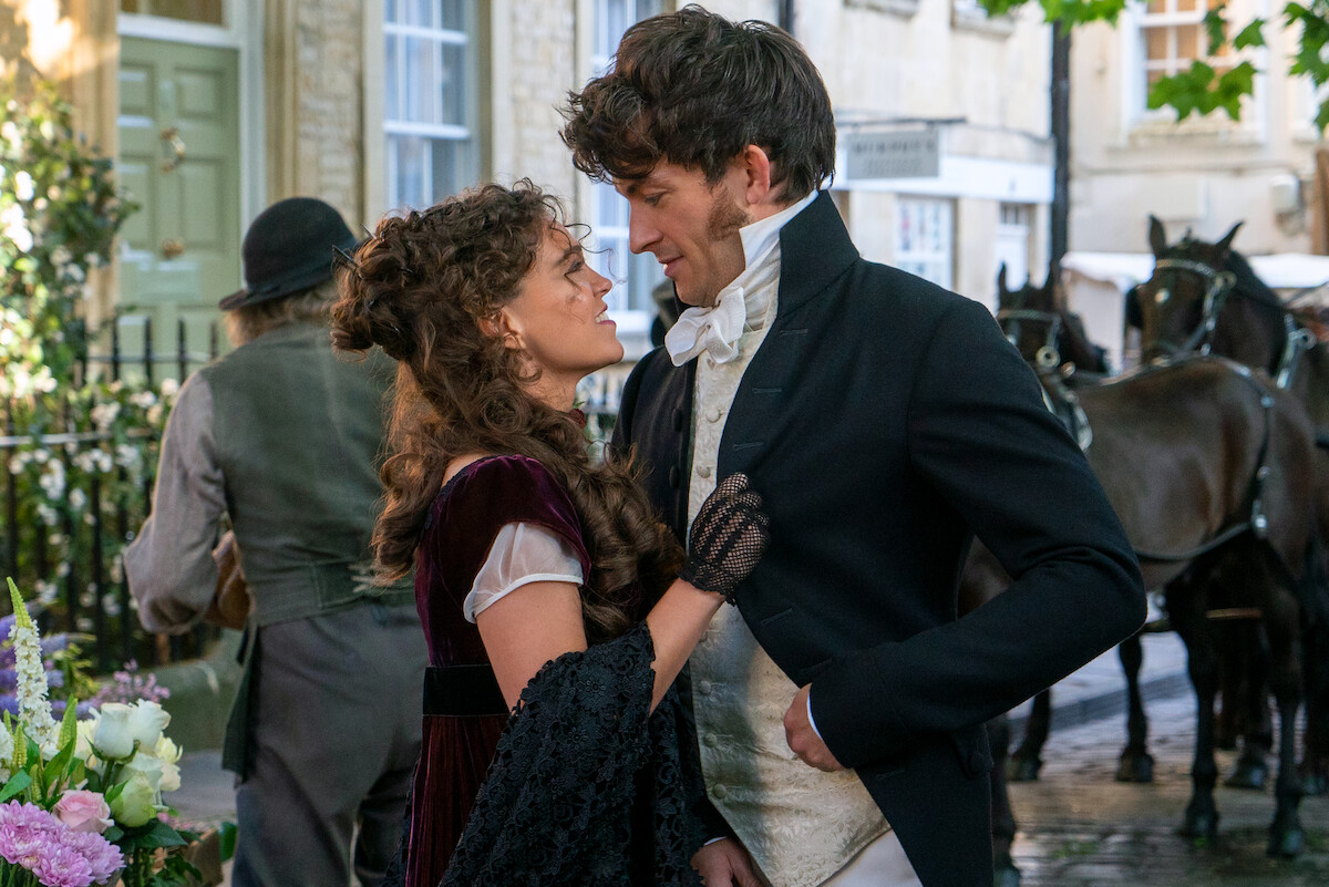 A woman and man in historical costumes gaze at each other on a cobblestone street with horses and carriages in the background, suggesting a romantic moment in a period setting.