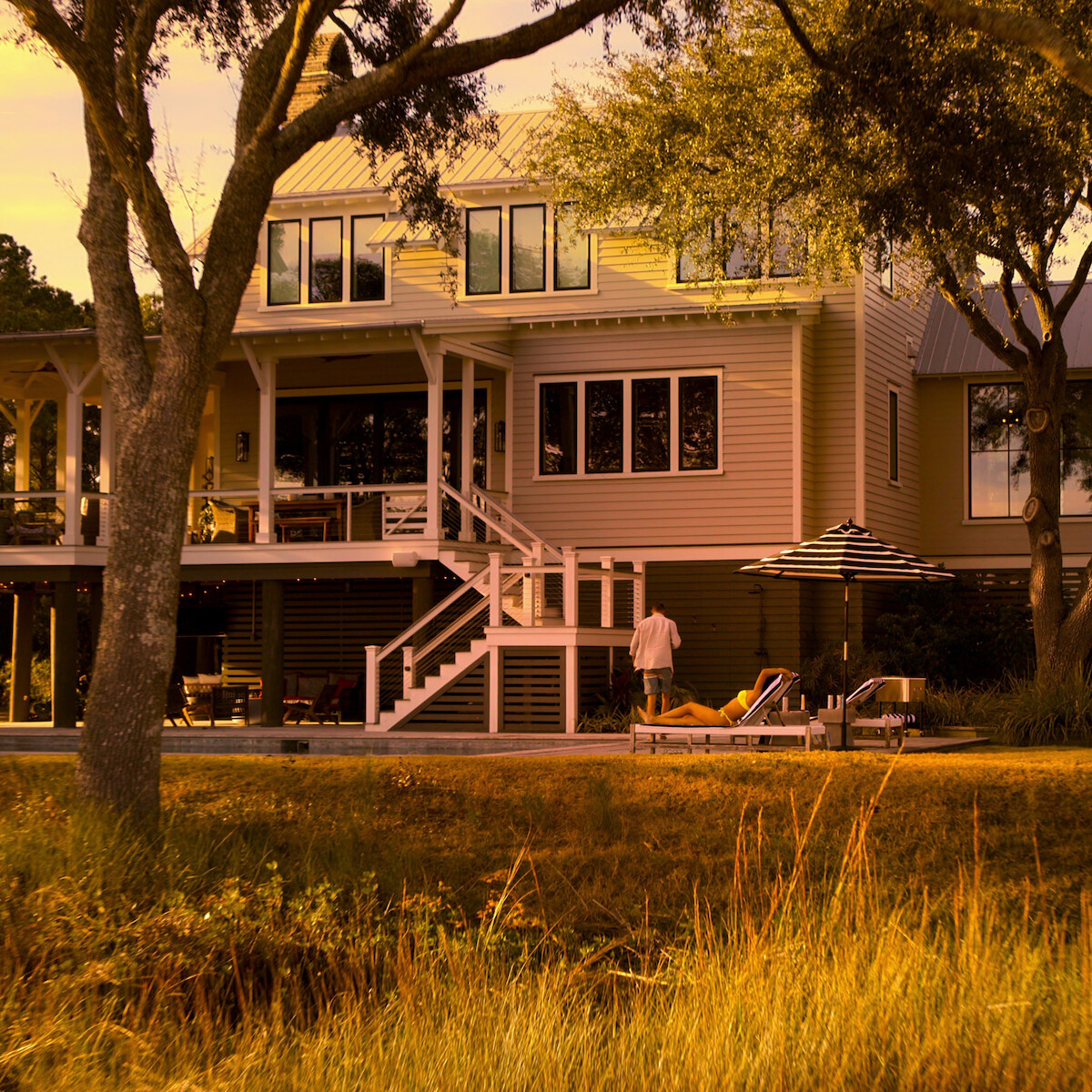 Modern house in a wooded setting at sunset, with trees in the foreground, a person standing by lounge chairs, another person relaxing under a striped umbrella on a patio. Warm lighting and tall grass in front.