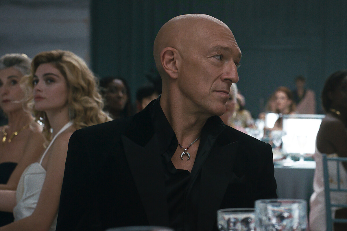 Bald man in a black suit sits at a formal event, surrounded by elegantly dressed people and decorated tables in a dimly lit banquet hall.