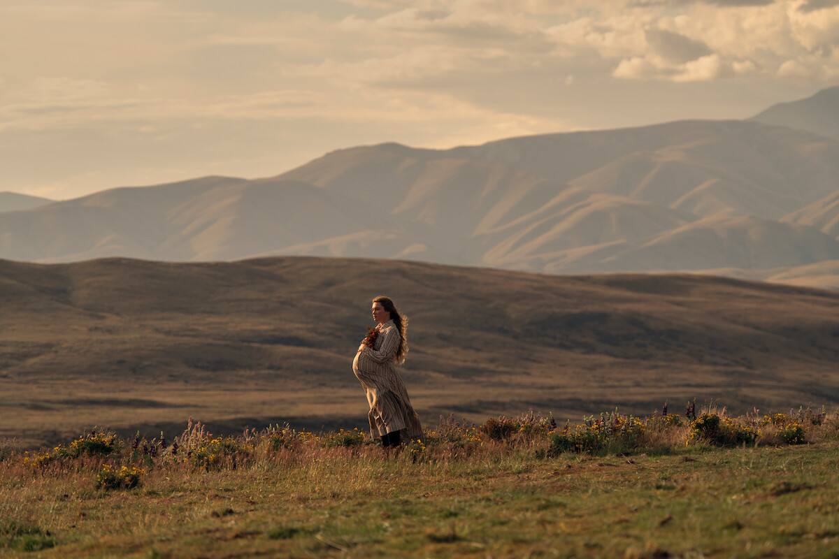 Pregnant woman standing in a grassy field at sunset, surrounded by wildflowers, with rolling hills and mountains in the background under a cloudy sky.