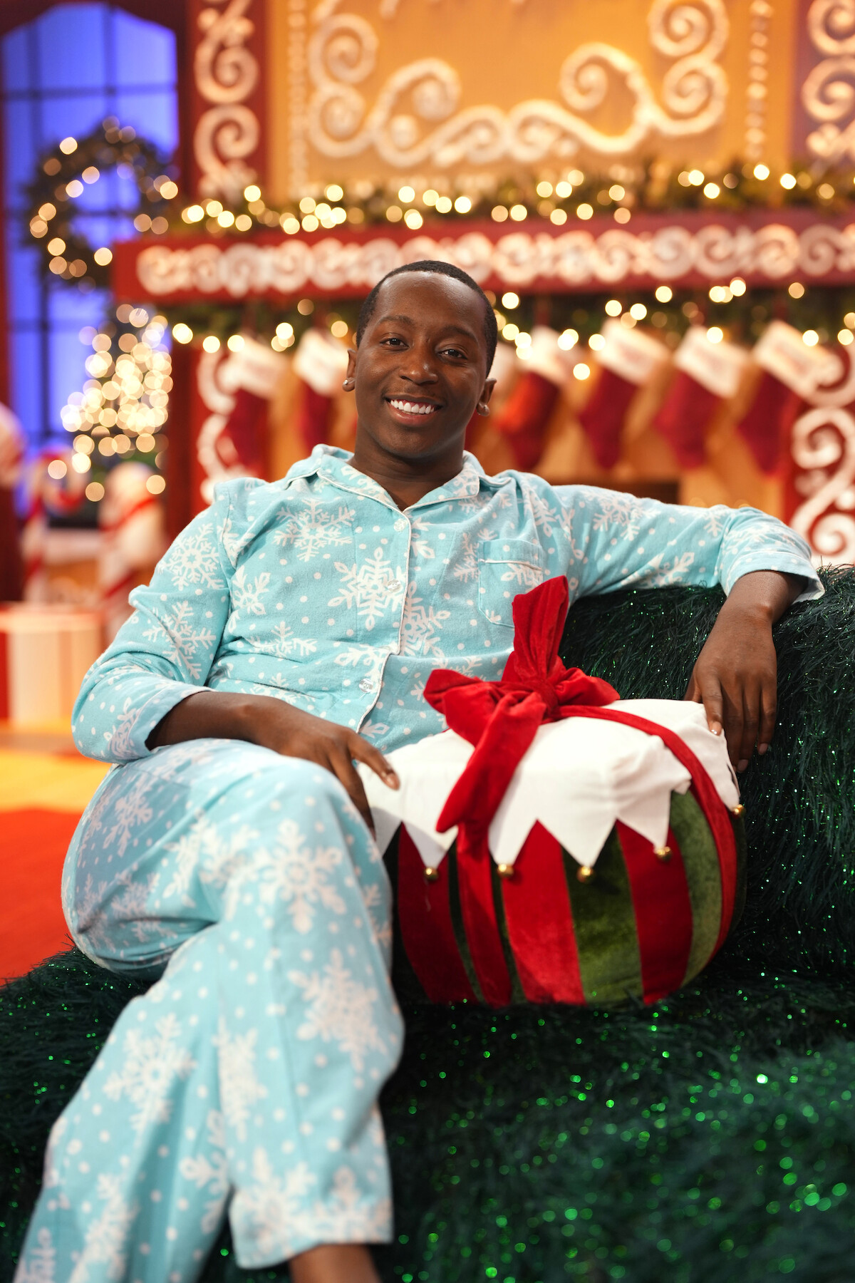 Person in blue pajamas sitting on a green couch holding a round Christmas gift, festive holiday background with stockings, garlands, and gingerbread-style decorations.