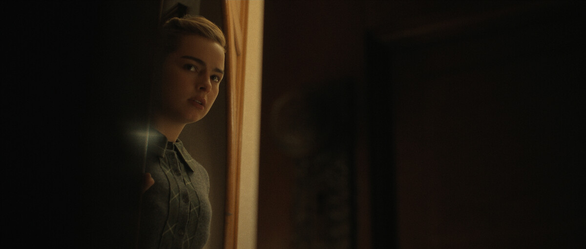 A young woman with a concerned expression peeks around a doorway into a dimly lit room.