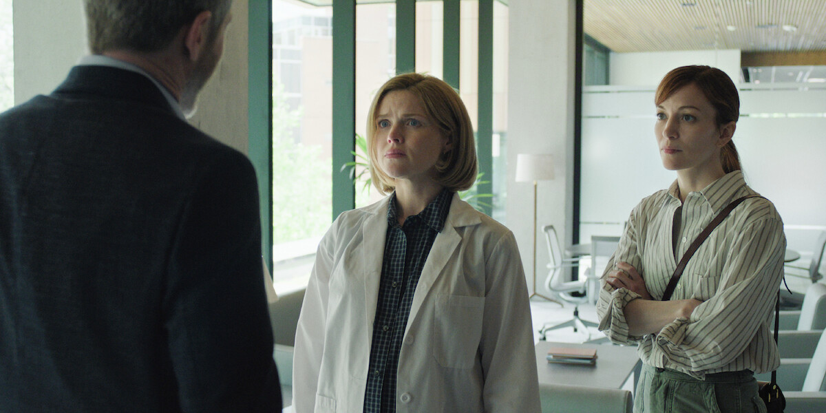 Two women, one in a lab coat and one in casual attire, stand in a modern office or lab with large windows, talking to a man whose back is to the camera.