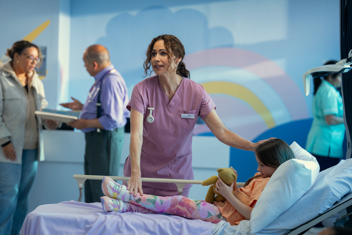 A nurse comforts a young girl in a hospital bed holding a stuffed animal in a colorful, child-friendly hospital room, while medical staff and a visitor interact in the background. The mood is warm and caring.