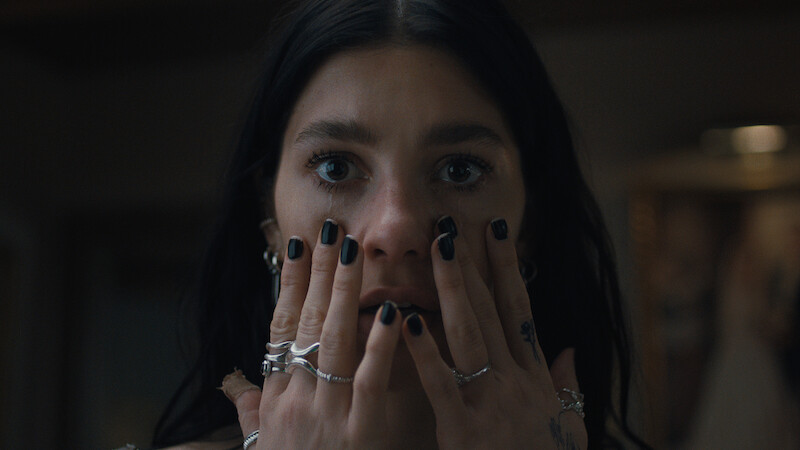 A woman with dark hair and black nail polish holds her hands to her face indoors, wearing multiple rings and looking surprised or concerned in a dimly lit setting.