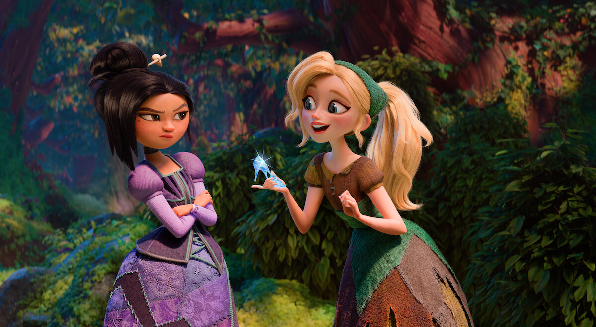 Two animated girls in medieval dresses stand in a lush forest. One girl is smiling and holding a glowing crystal, while the other looks skeptical with arms crossed. Sunlight filters through the trees in the background.