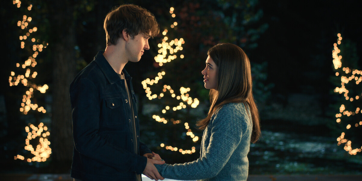 A young man and woman stand outdoors at night, holding hands and looking at each other, with trees decorated in glowing lights in the background.