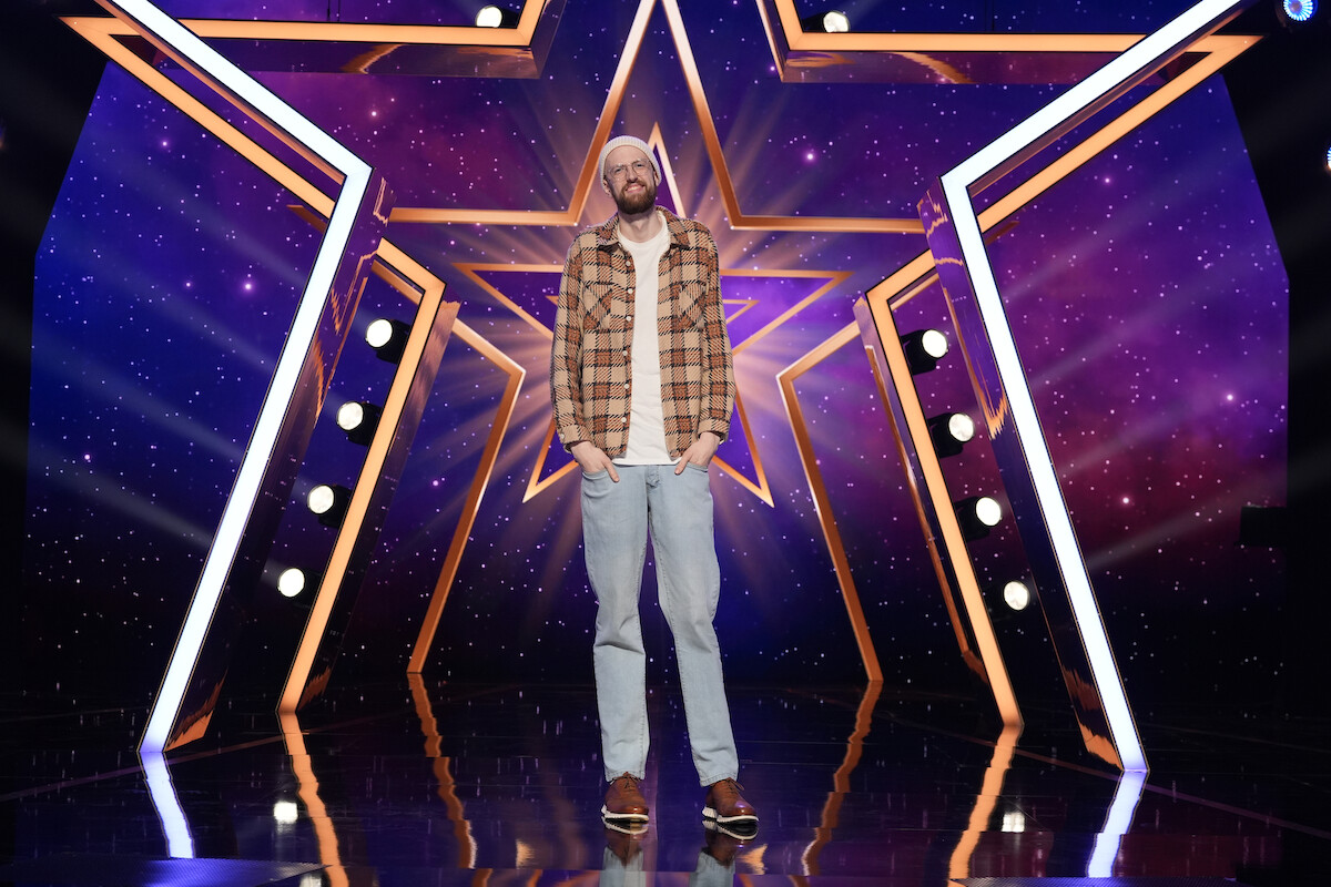 Liam Nelson in casual clothes stands on a stage with a large illuminated star backdrop and spotlights, with a vibrant purple and blue galaxy-themed background.