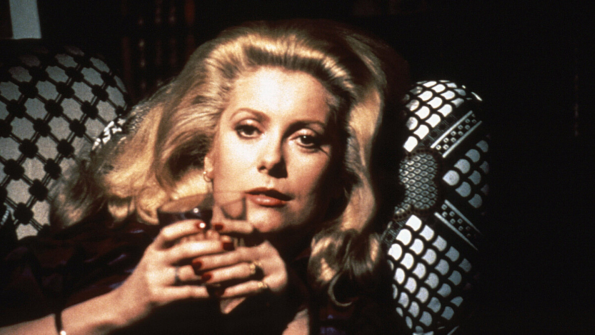 Woman with blonde hair sitting in a dimly lit room, holding a glass with both hands, looking directly at the camera, patterned chair in the background, intimate and contemplative mood.