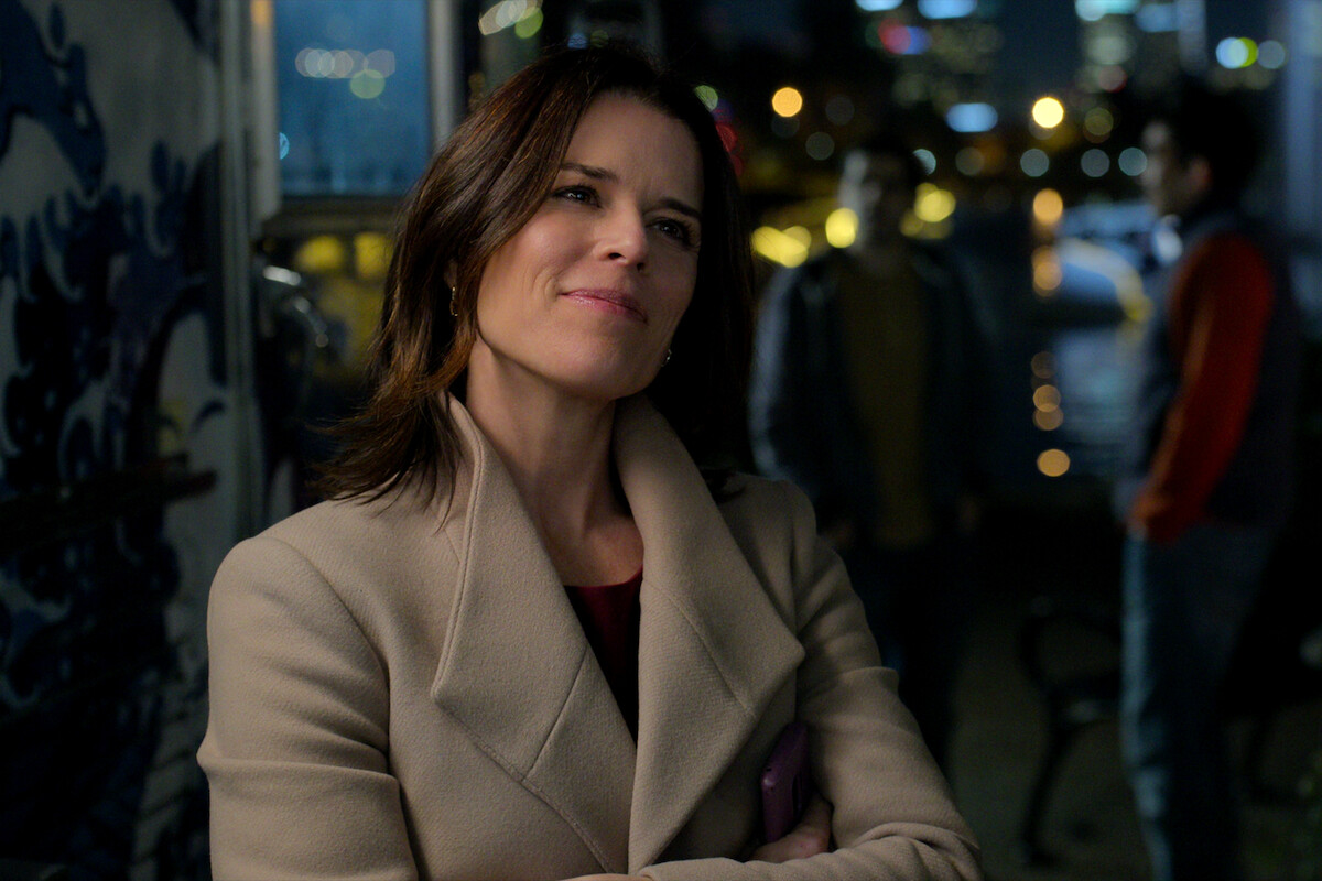 Woman in a beige coat smiling with arms crossed, standing outdoors at night with blurred city lights and three people in the background.
