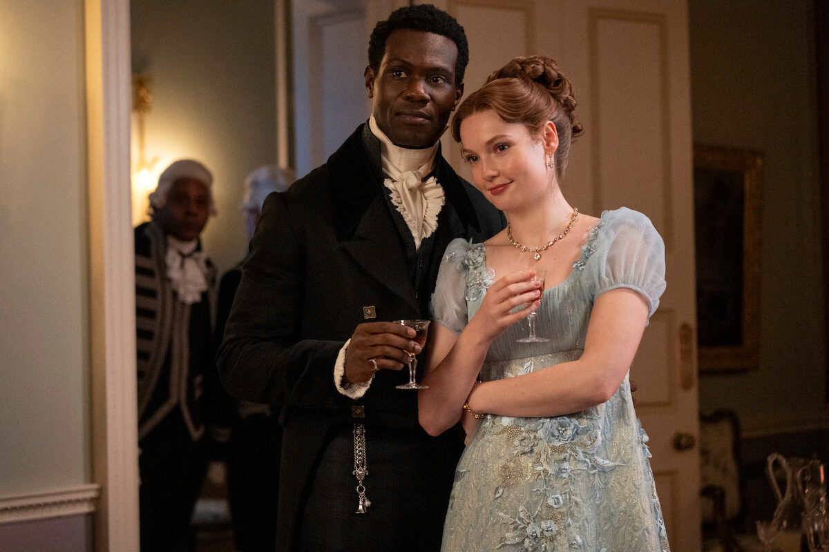 A well-dressed man and woman in Regency-era attire stand closely together at an elegant indoor gathering, each holding a drink, with soft lighting and classical decor in the background.