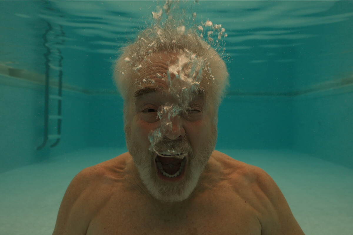 Elderly man underwater in a swimming pool, exhaling bubbles, with a ladder and pool walls visible in the background, creating a dynamic and playful underwater scene.