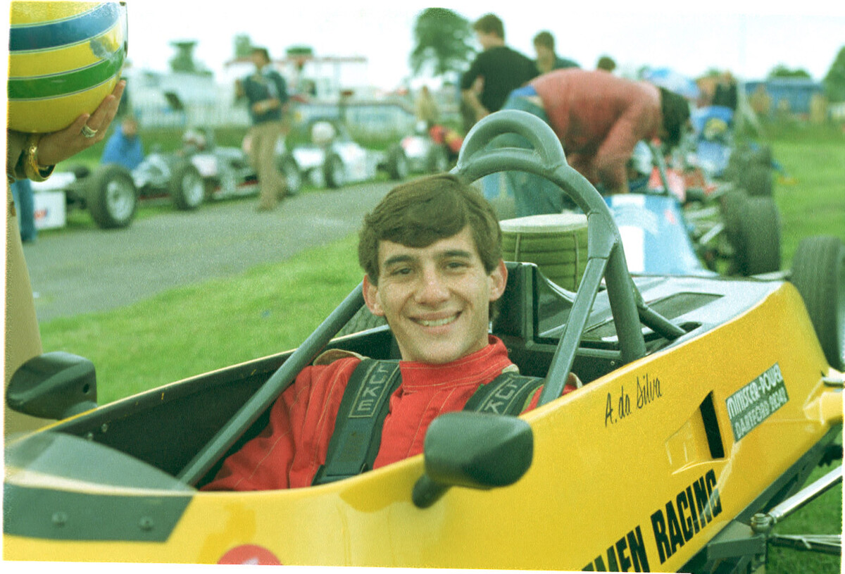 A teenaged Ayrton Senna wears a red shirt and sits in a yellow car, smiling adorably!