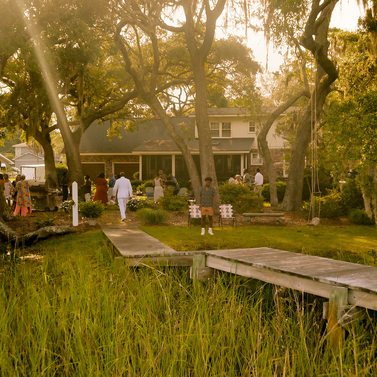 People gather in the backyard of a house surrounded by large trees and greenery, with sunlight streaming in. A dock leads from the marsh toward the gathering, evoking a relaxed outdoor event or family gathering in a natural setting.