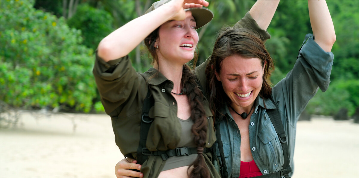 Two women in outdoor clothing, one comforting the other, both emotional on a sandy beach with lush green trees in the background, suggesting a survival or adventure scenario in a natural tropical setting.