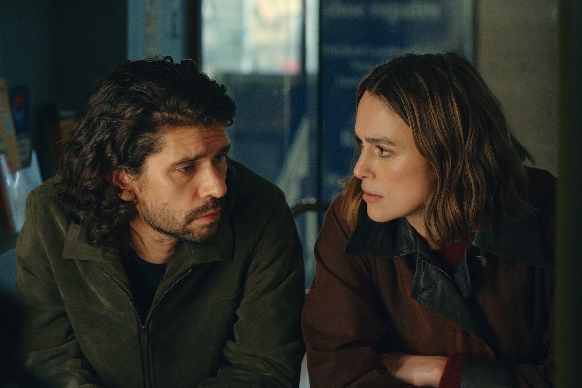 Two people sit closely at a table indoors, having a serious conversation. Both wear jackets and appear focused on each other, with a blurred background suggesting a casual or café setting.