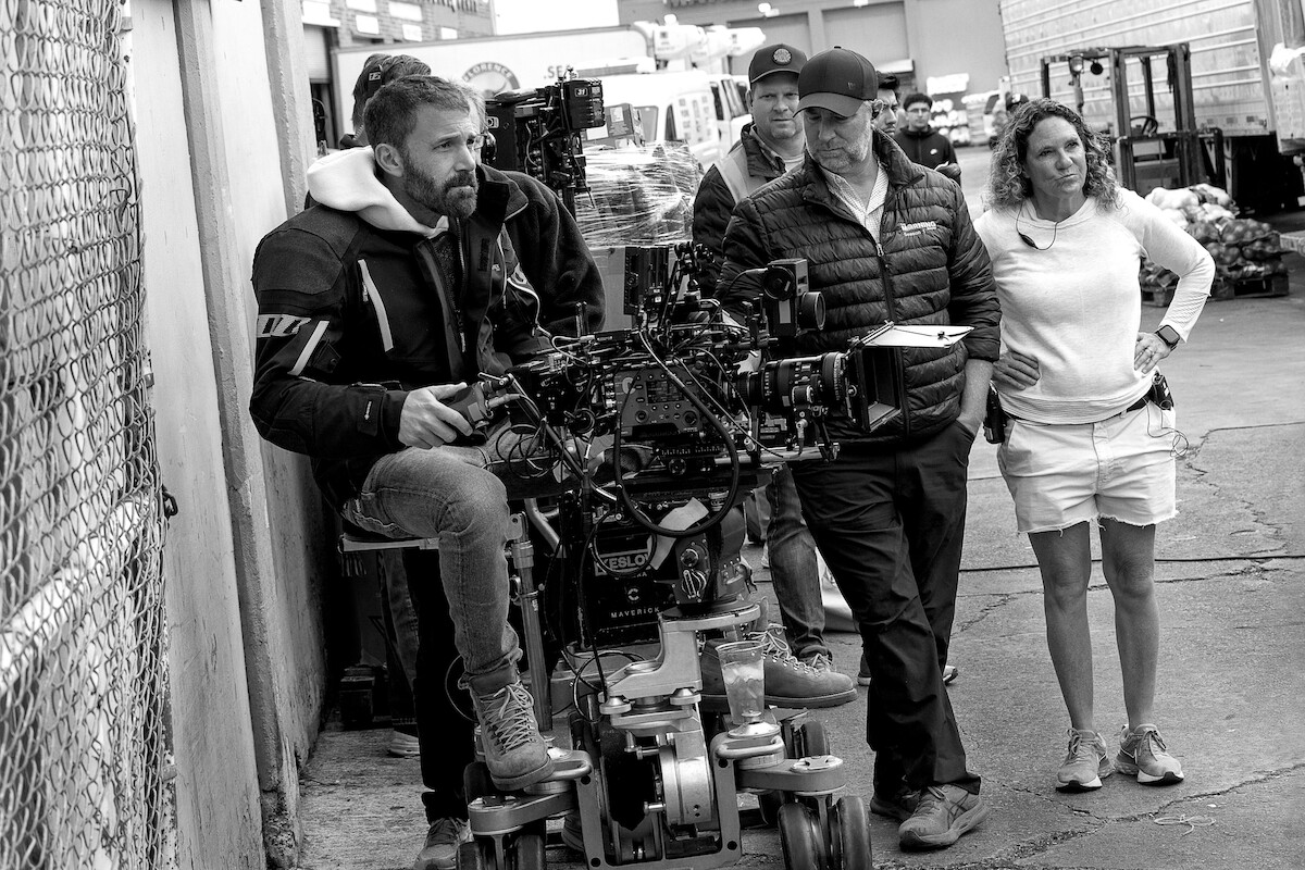 Film crew on outdoor set, including a cameraman operating camera equipment on a dolly, surrounded by crew members in casual attire near a chain-link fence and industrial buildings.