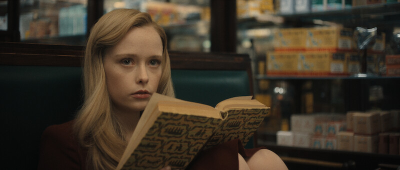 A young woman with long blonde hair sits in a booth, holding an open book and staring ahead with a serious expression in a store or diner setting.