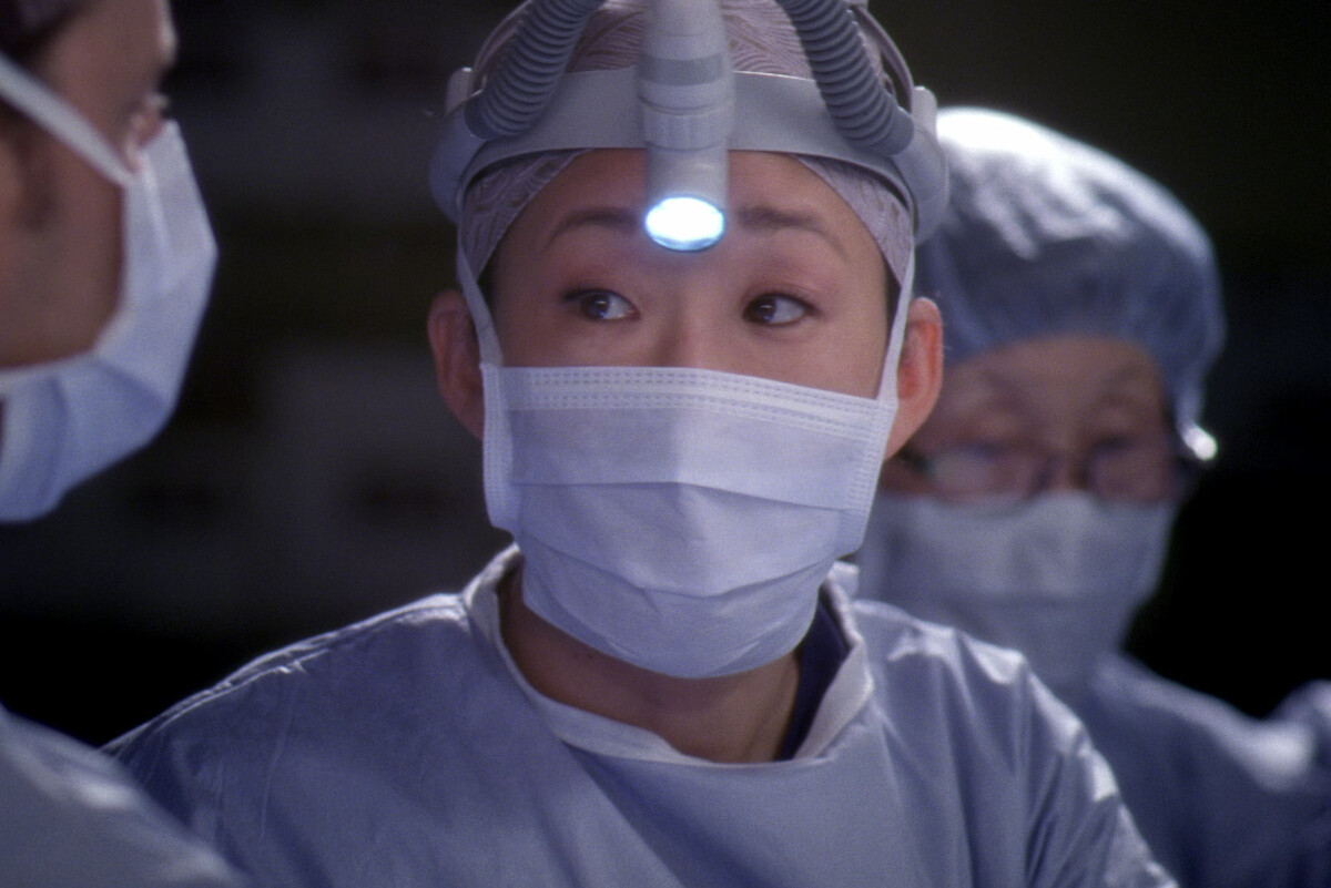 Sandra Oh in full scrubs, wearing a headlamp, looks at another doctor. 