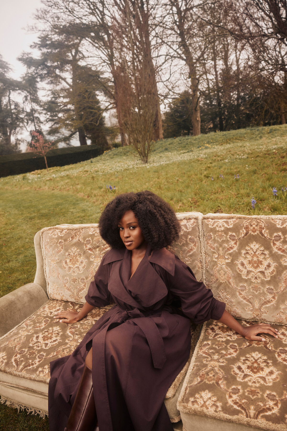 Woman in a brown coat sitting on an ornate vintage couch outdoors in a grassy garden with trees and overcast sky in the background.