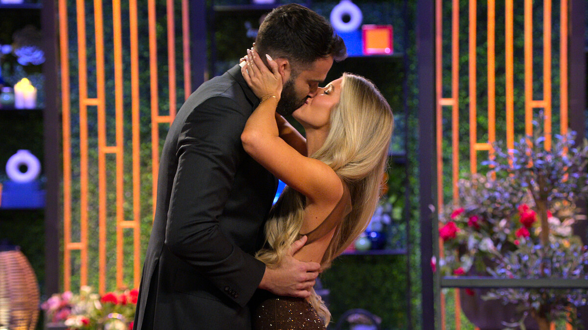 Alex Henderson and Ashley Carpenter share a kiss indoors, surrounded by decorative plants, modern lighting, and a colorful, elegant setting.