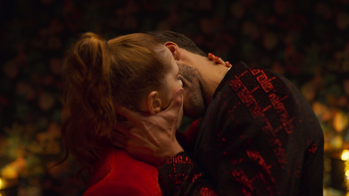Two people passionately kissing in a dimly lit, intimate setting with floral wallpaper in the background and warm, ambient lighting.