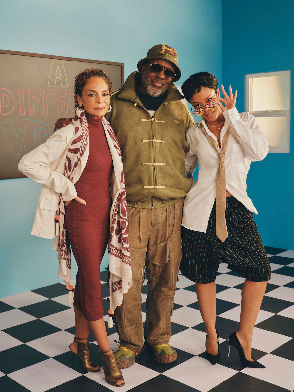 Three people stand together on a black-and-white checkered floor in a colorful room with blue walls and a window, next to a chalkboard that reads "A Different World." Each person is dressed in stylish, distinct clothing.