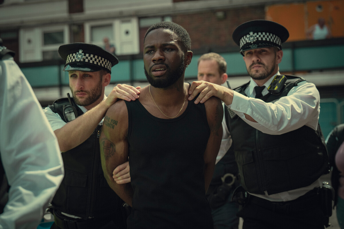 Joshua Blisset as Kieron stands with his hands behind his back as police officers detain him in Season 3 of ‘Top Boy.’