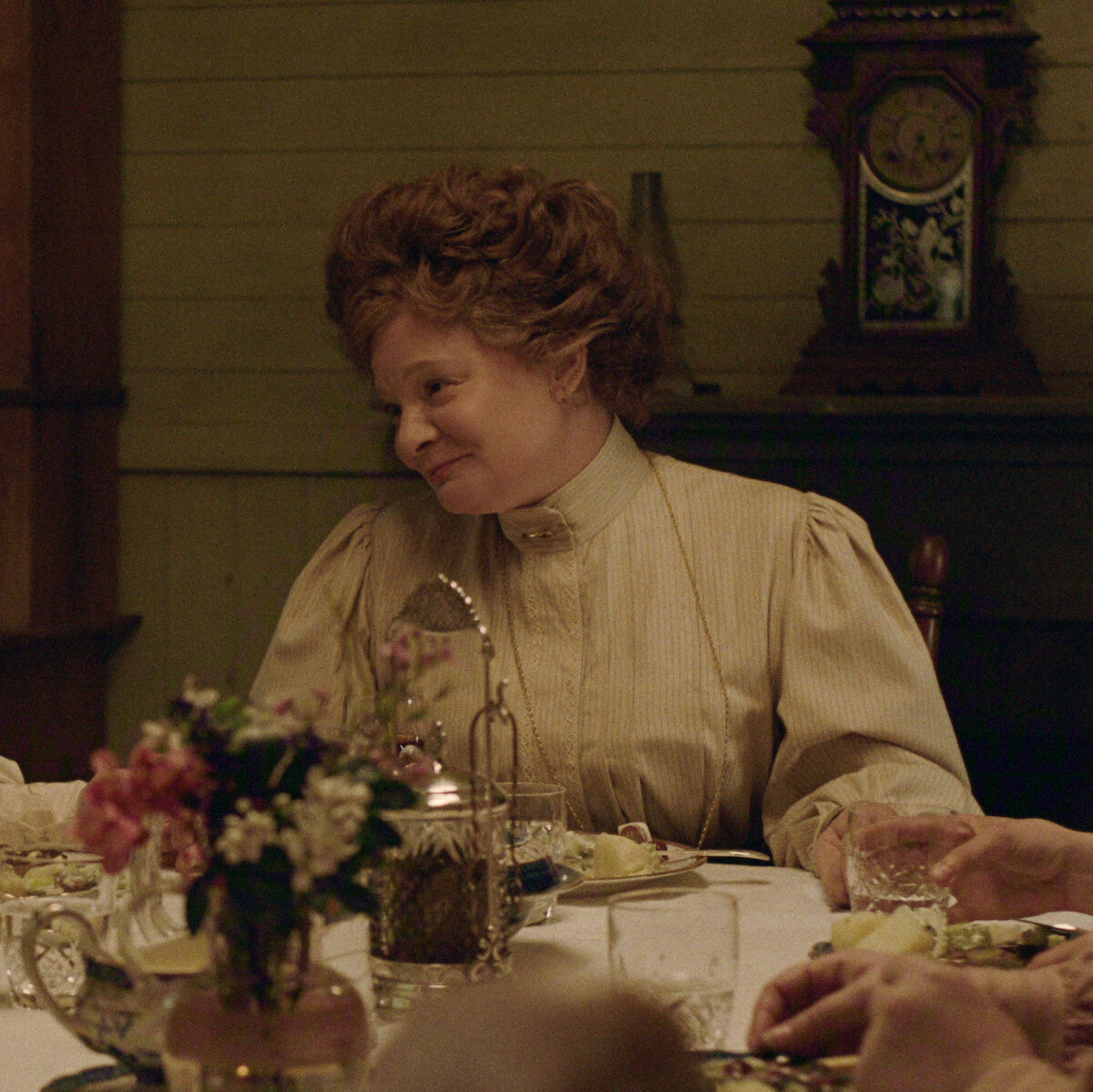 Martha Plimpton as Faye in vintage clothing sits at a dinner table set with flowers and dishes in a warmly lit, old-fashioned room with a decorative clock in the background.