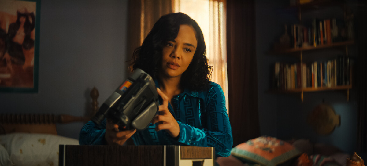 A woman in a blue shirt examines a handheld video camera in a cozy, softly lit bedroom with bookshelves, a bed, pillows, and warm sunlight filtering through the curtains.