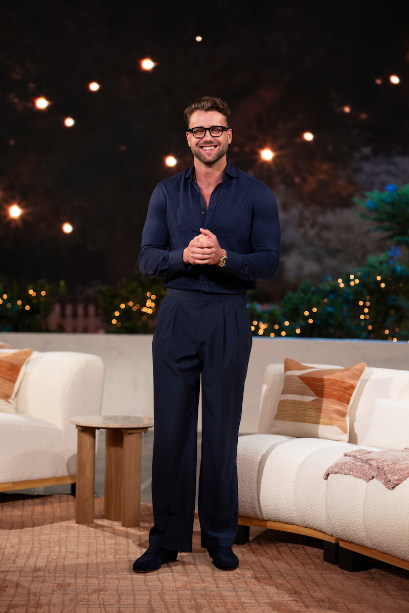 Smiling man in dark clothes stands between white couches in a cozy, warmly lit living room with string lights and greenery in the background, creating a relaxed, inviting atmosphere.