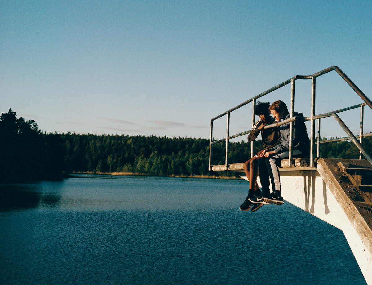 Yassir Hassan as Billy and Olle Strand as Dogge sit together on a diving board over a lake in ‘Deliver Me.’