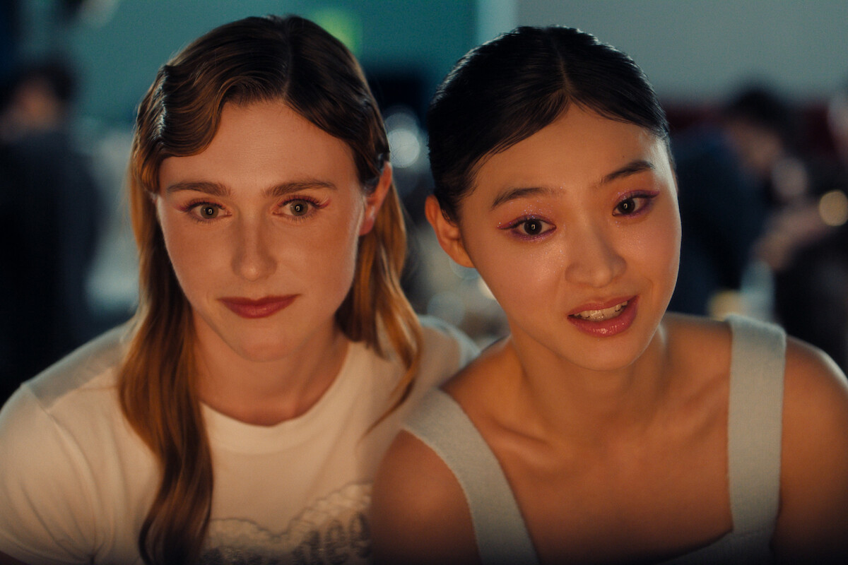 Two young women sit closely together indoors at an event, smiling softly at the camera with a blurred background suggesting a lively, social atmosphere.
