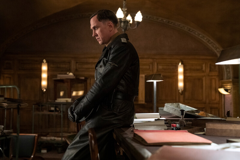 Lars Eidinger as Sergeant Major Reinhold von Rumpel sitting on the edge of a desk, looking down. 