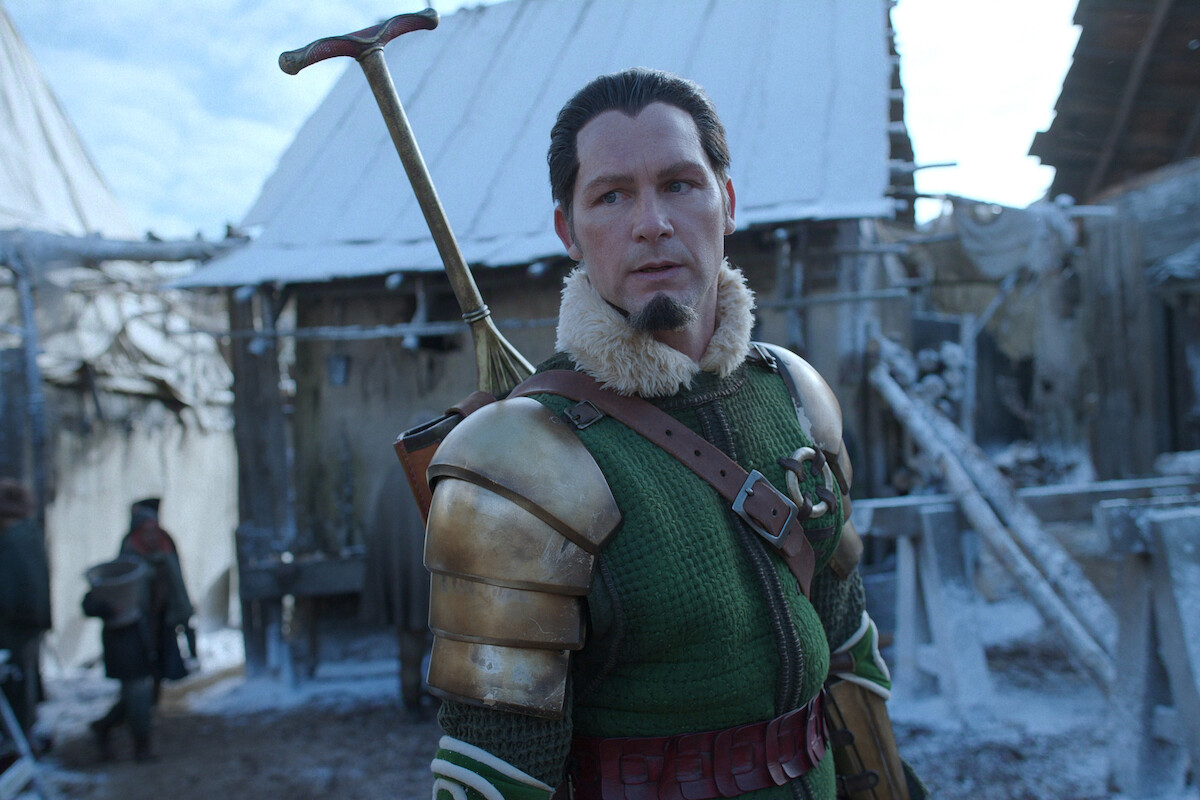 A man in green medieval armor with gold pauldrons and a fur collar stands outdoors in a snowy village, carrying a weapon on his back; rustic wooden buildings and snow-covered ground are visible in the background.
