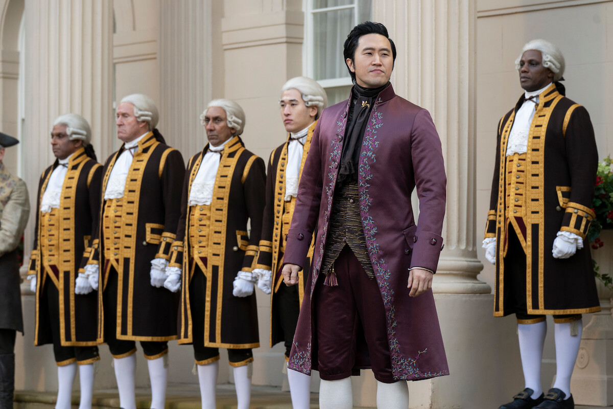A man in an ornate maroon 18th-century costume stands confidently in front of uniformed footmen in formal wigs and black-and-gold attire, with classical columns of an elegant building in the background.