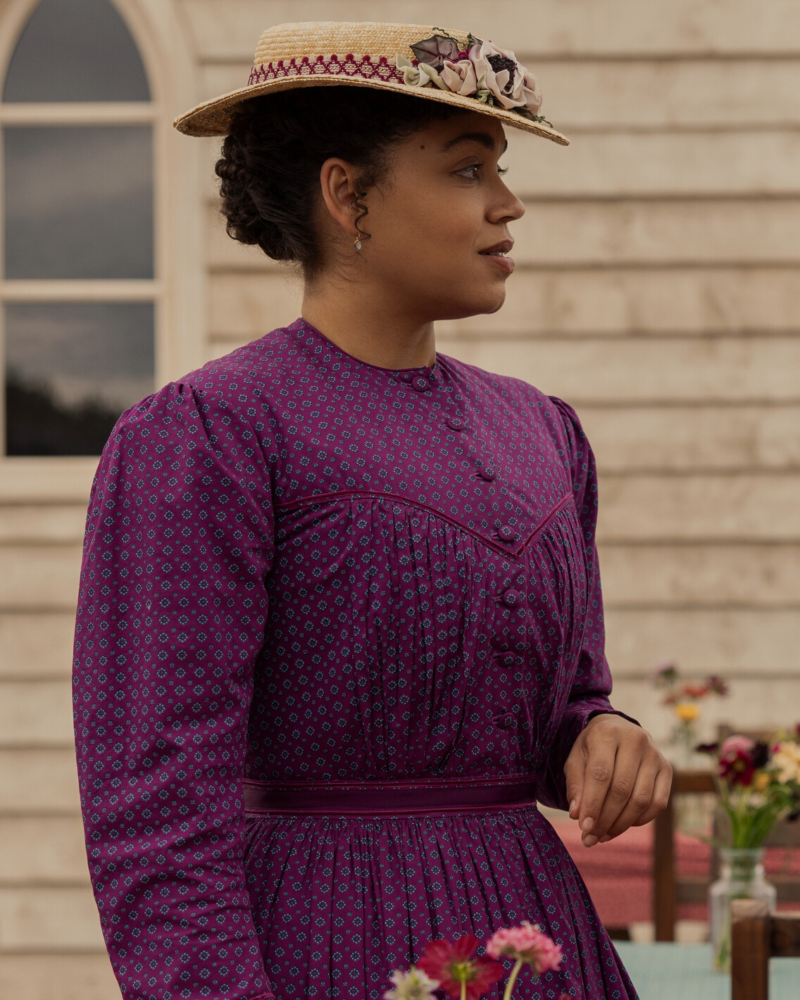 Woman in a purple patterned dress and straw hat with flowers, standing outdoors by a wooden building, with a window and flowers in the background, looking to the side in daylight.