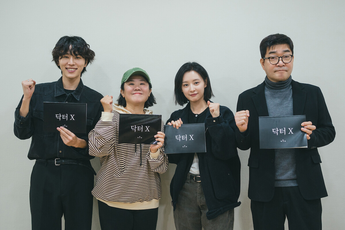 Quatro pessoas asiáticas posam para uma foto com um cartaz nas mãos, no qual se lê “Dr. X” escrito em coreano