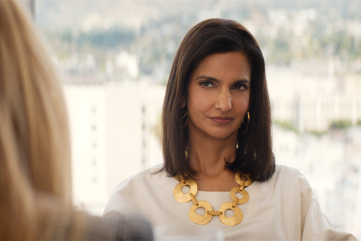 Woman with straight dark hair and a bold gold necklace sitting indoors, wearing a white blouse, looking to the side with a slight smile; blurred cityscape and buildings visible through a window in the background.