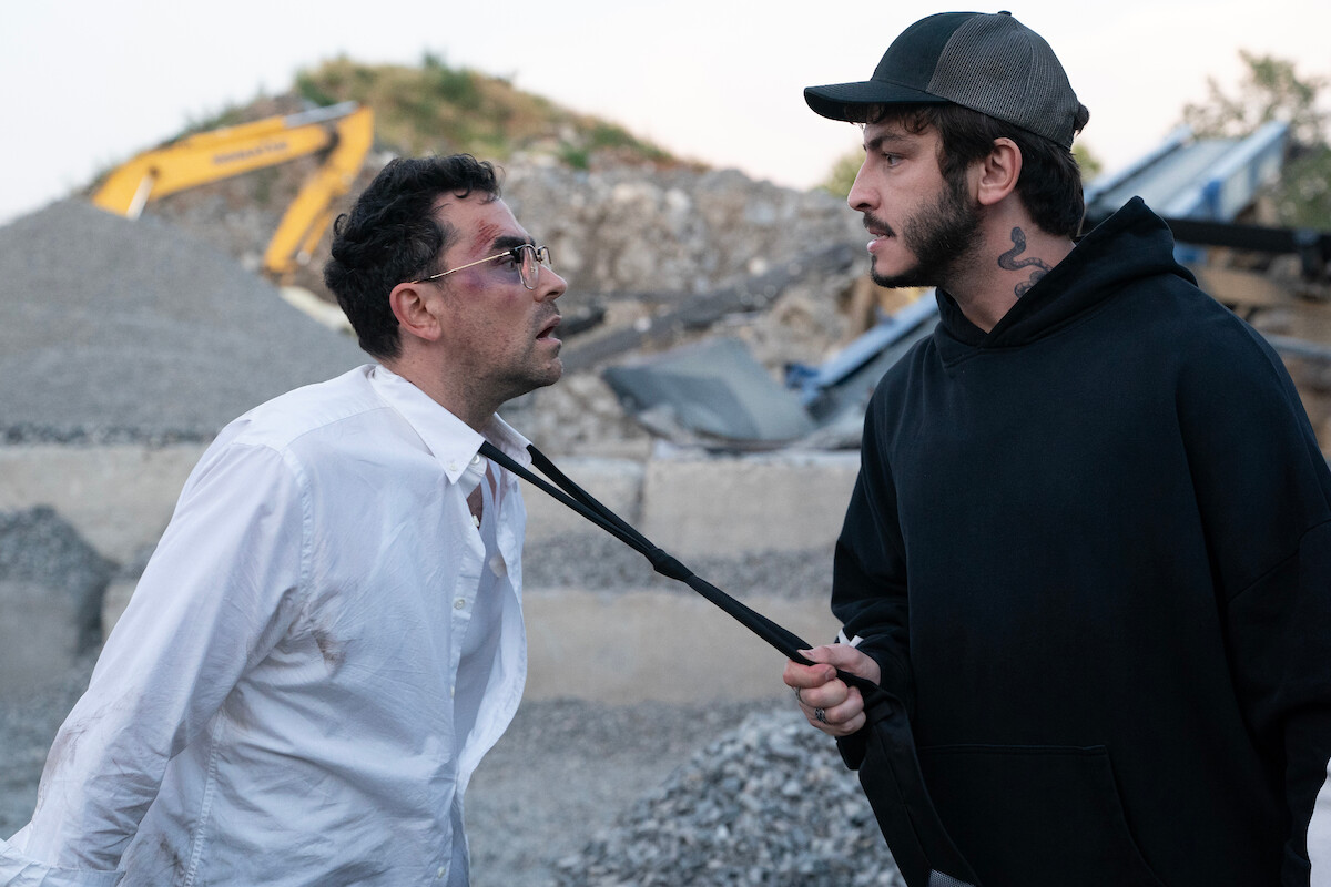 Two men confront each other at a construction site; one, in a white shirt with a bruised face, is being grabbed by the collar by the other, who wears a black hoodie and cap. Dirt piles and machinery are visible in the background.