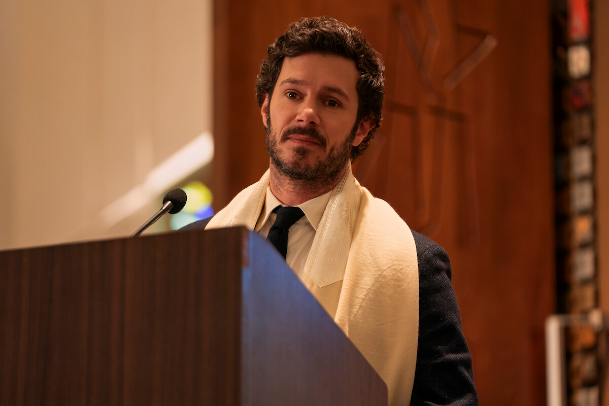 Man wearing suit and tallit standing at a podium with a microphone, speaking in a synagogue or religious setting with wood paneling and soft lighting in the background.