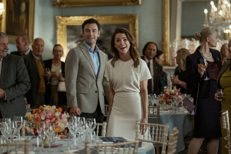 A man in a light suit and a woman in a white dress stand smiling and looking ahead at a formal event, surrounded by guests holding drinks in an elegantly decorated room.