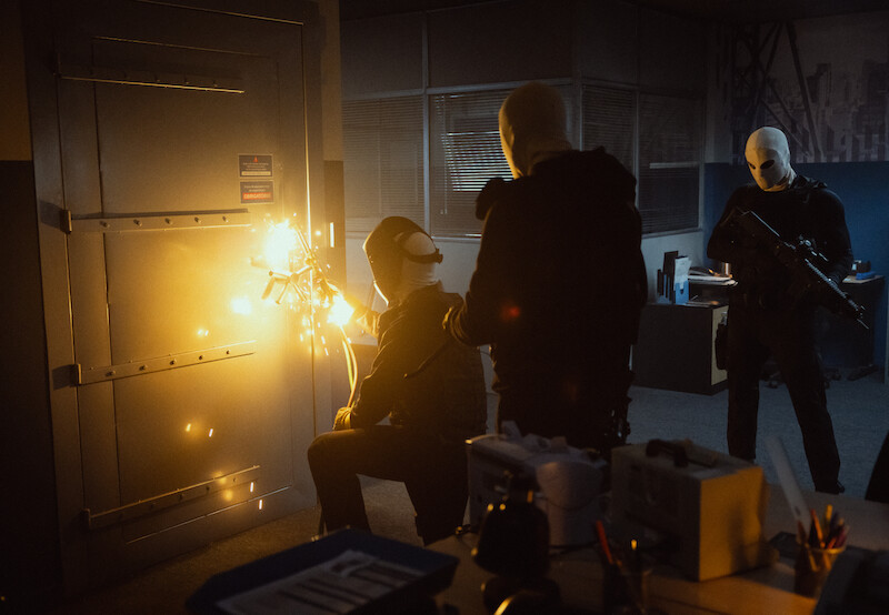 Three masked individuals in dark clothing break into a secure room using a blowtorch, sparks flying, while one stands guard with a rifle in an office setting.




