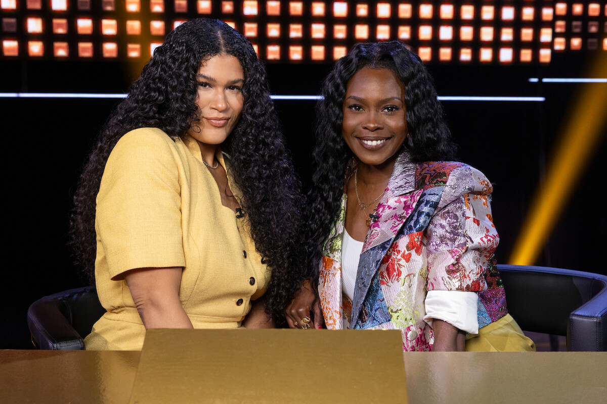 Two women sit side by side at a table on a brightly lit stage, smiling. One wears a yellow outfit, the other a colorful jacket. The background has a grid of illuminated squares, creating a lively, upbeat atmosphere.