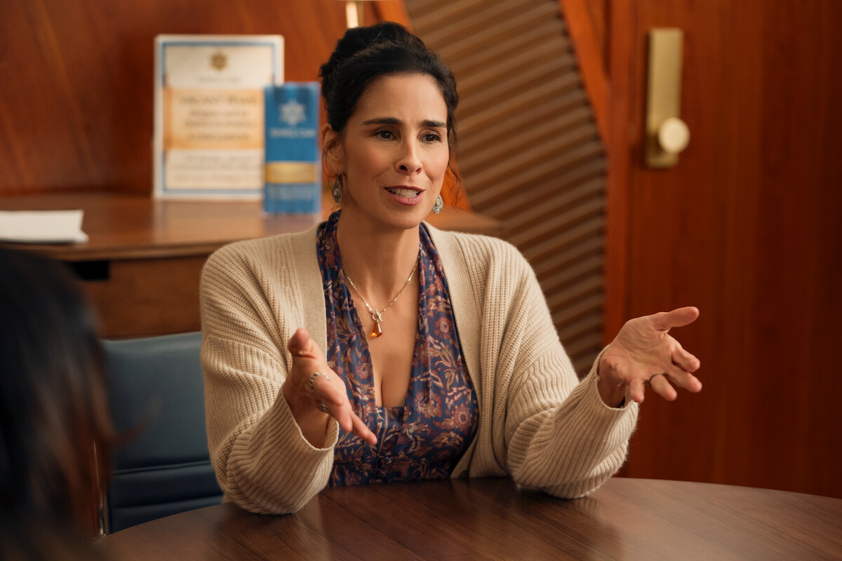 Woman with dark hair in a bun, wearing a patterned dress and cream cardigan, sits at a wooden table in an office, gesturing as she speaks, with certificates and a blue book visible on a shelf behind her.