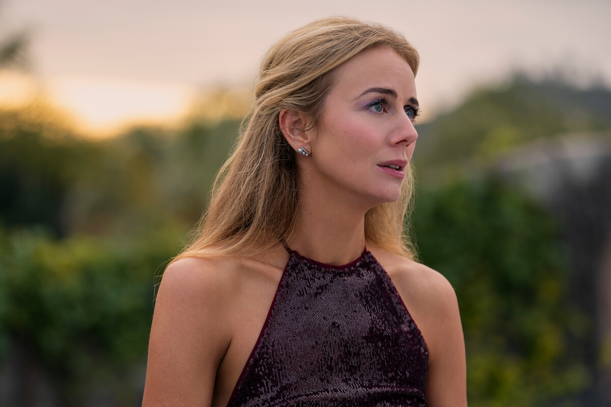Woman with long blonde hair wearing a sleeveless, sequined burgundy dress stands outdoors at sunset, surrounded by greenery, looking thoughtfully to the side.