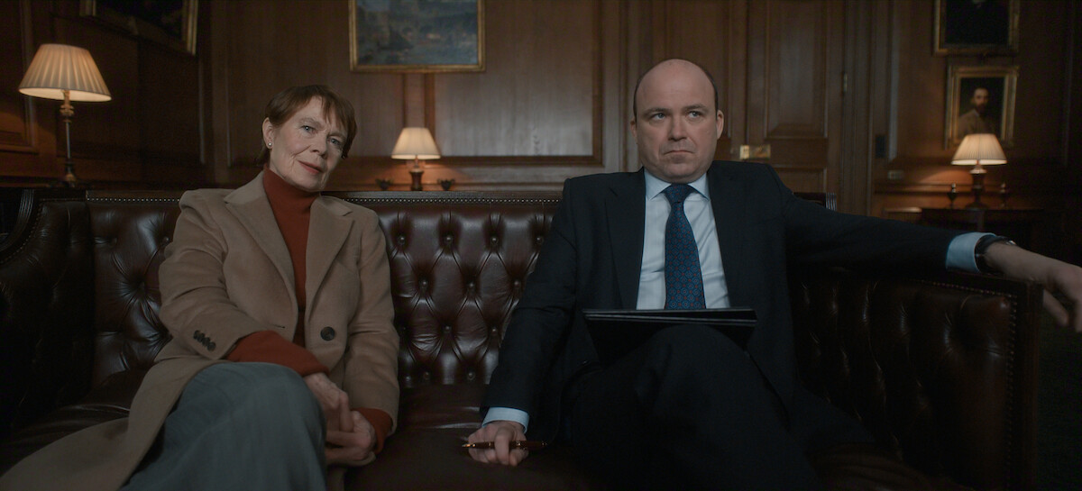 Two people sit on a brown leather sofa in a wood-paneled office with warm lighting, lamps, and framed art. The woman wears a tan coat and red turtleneck, and the man wears a suit, holding a pen and a folder, both with serious expressions.
