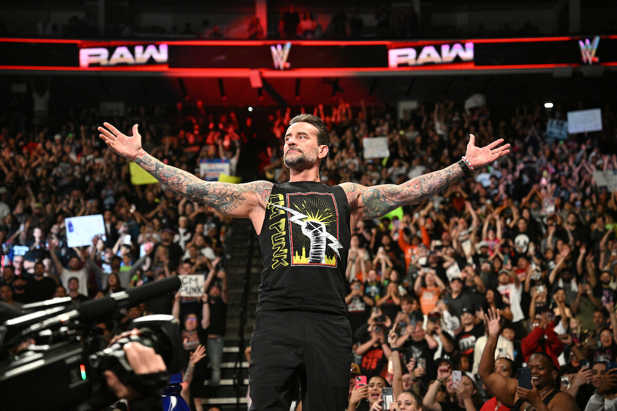 CM Punk with arms outstretched stands confidently in a packed arena during a WWE Raw event, with a large, cheering audience and bright lights in the background.