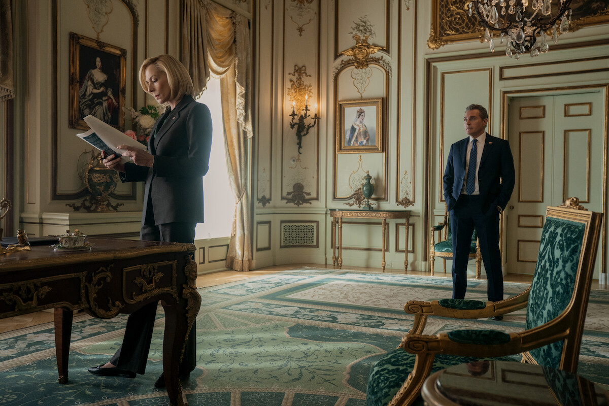 A woman in a business suit reads papers at a desk while a man in a suit stands nearby in an elegant, ornate room with portraits, chandeliers, and luxurious decor.